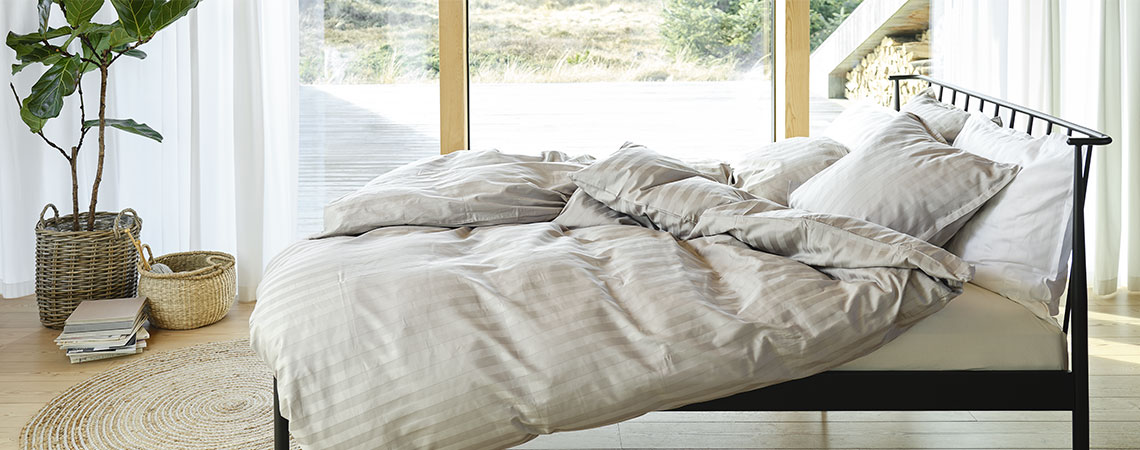 Dormitor cu pat metalic negru, pilote și perne, acoperit cu lenjerie de pat cu dungi de culoare gri deschis și albe