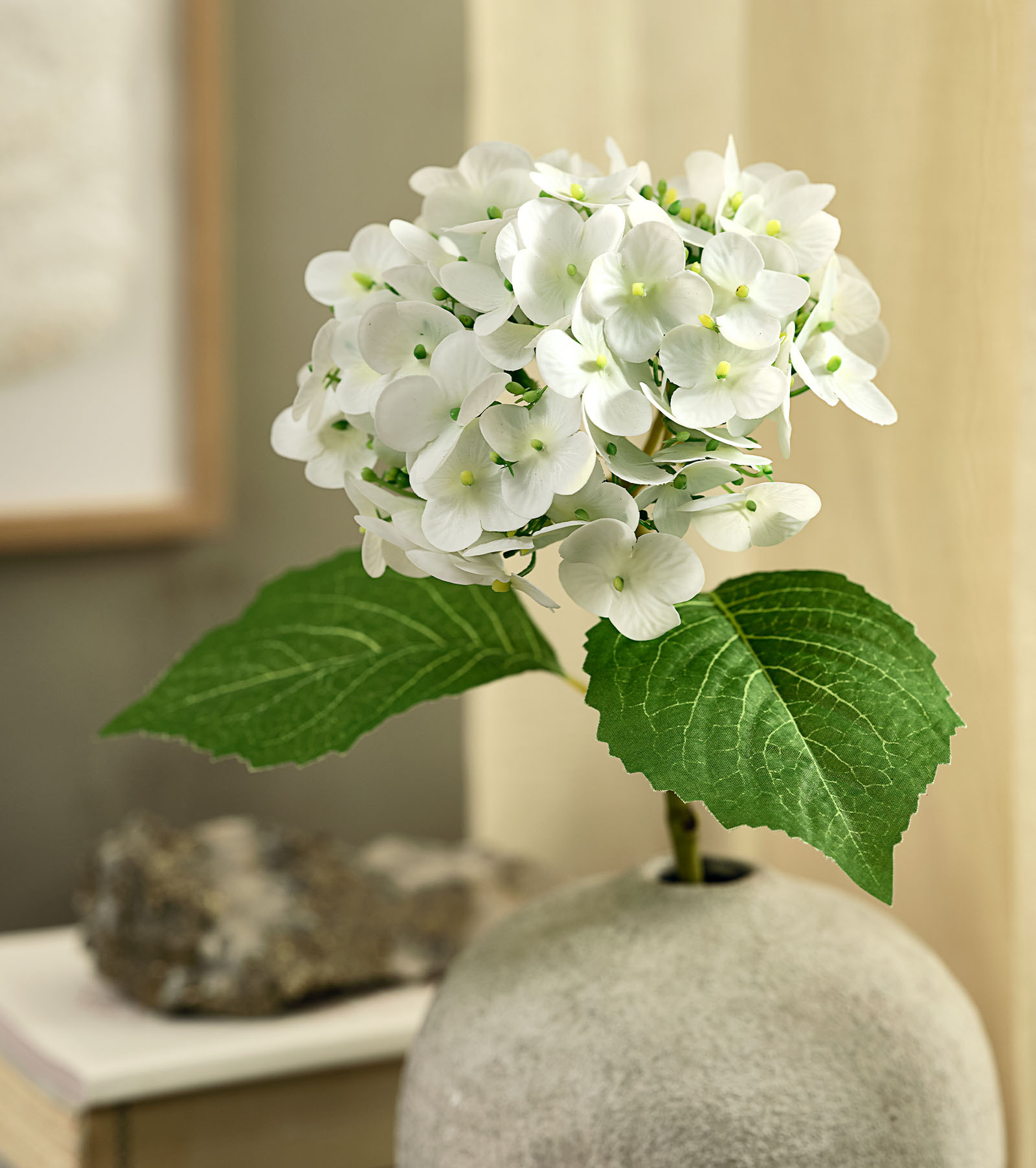 Floare artificială cu aspect de hortensie albă cu frunze verzi într-o vază rustică
