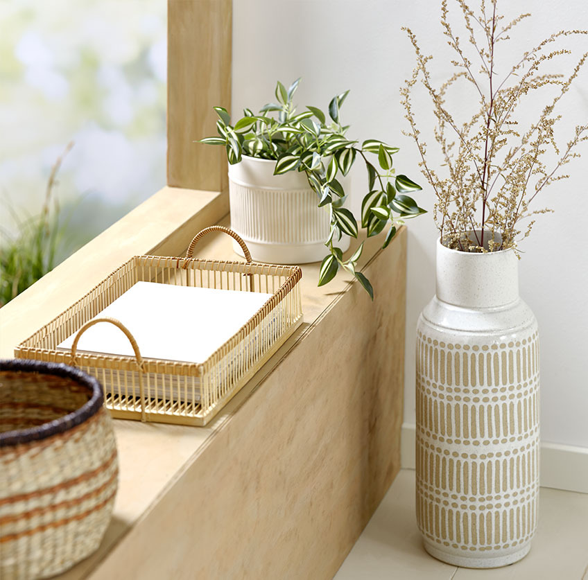 Vază înaltă lângă pervaz cu tavă din bambus și ghiveci alb cu plantă artificială